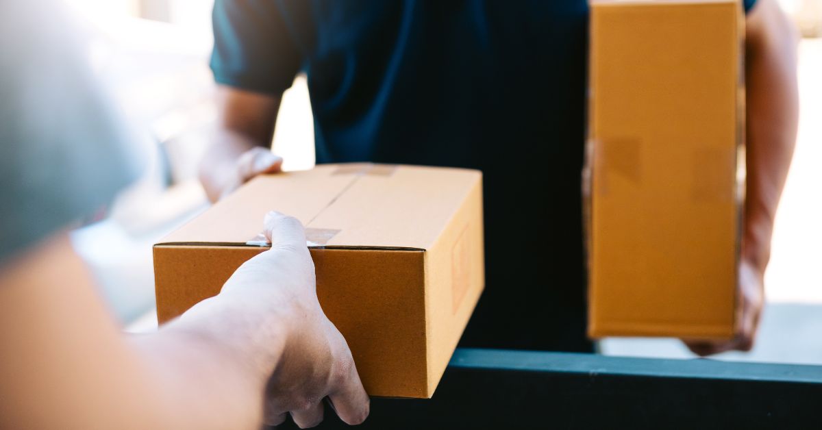 Entregador entrega uma caixa de papelão a um cliente na entrada de um imóvel, enquanto segura outra encomenda. A imagem destaca o momento da entrega, com foco nas caixas e nas mãos das pessoas, e o fundo desfocado.