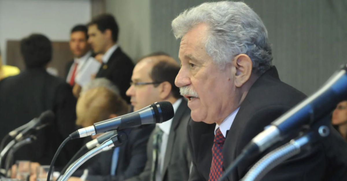 Homem idoso de cabelos grisalhos e bigode branco fala ao microfone durante uma reunião ou audiência pública. Vestindo terno escuro, camisa branca e gravata vermelha com estampa discreta, ele aparece em primeiro plano, de perfil, enquanto outras pessoas também estão sentadas à mesa ao fundo, em ambiente interno.