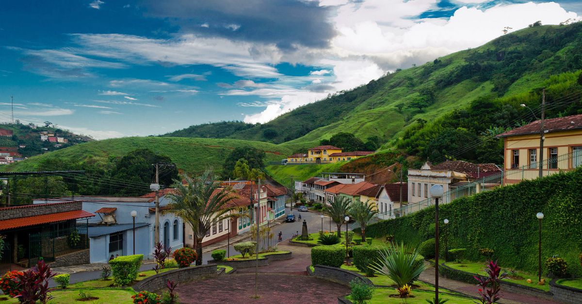 Vista panorâmica da cidade de Piau, na Zona da Mata mineira, com praça arborizada em primeiro plano, casas ao redor e morros verdes ao fundo sob um céu parcialmente nublado.