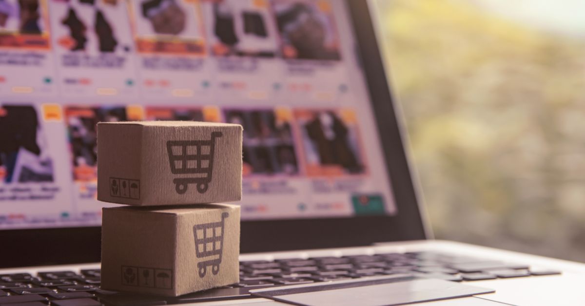 Em primeiro plano, duas pequenas caixas de papelão empilhadas sobre o teclado de um notebook. Nas laterais das caixas há o desenho de um carrinho de compras, símbolo associado ao comércio eletrônico. Ao fundo, desfocado, aparece a tela do computador exibindo uma página com diversos produtos organizados em grade, semelhante a uma loja virtual.