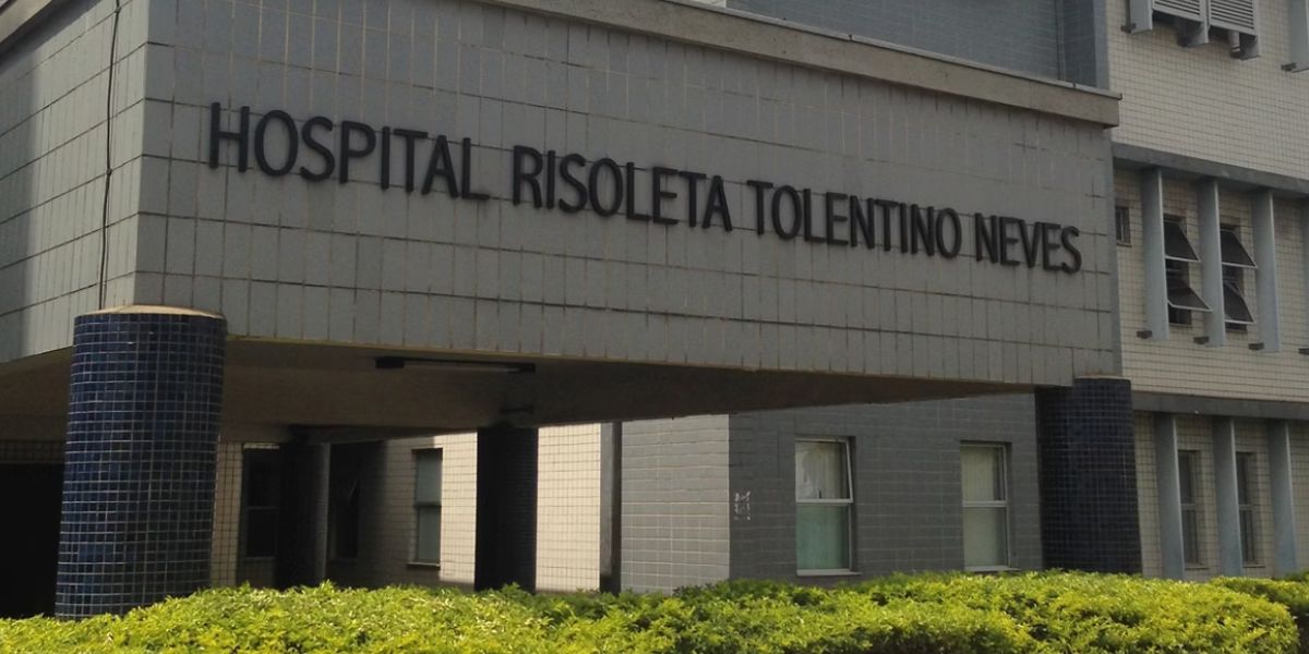 Fachada do Hospital Risoleta Tolentino Neves, em Belo Horizonte, com o nome da instituição em letras pretas na entrada do edifício. O prédio possui revestimento em tons de cinza, colunas escuras e área ajardinada com arbustos na parte frontal.