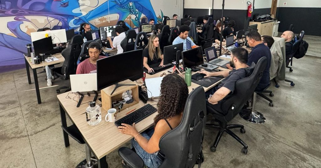 Fotografia colorida de um escritório moderno em conceito aberto. Cerca de quinze pessoas trabalham sentadas em mesas compartilhadas, organizadas em fileiras. Cada profissional utiliza computadores com monitores grandes, notebooks e outros equipamentos de trabalho.