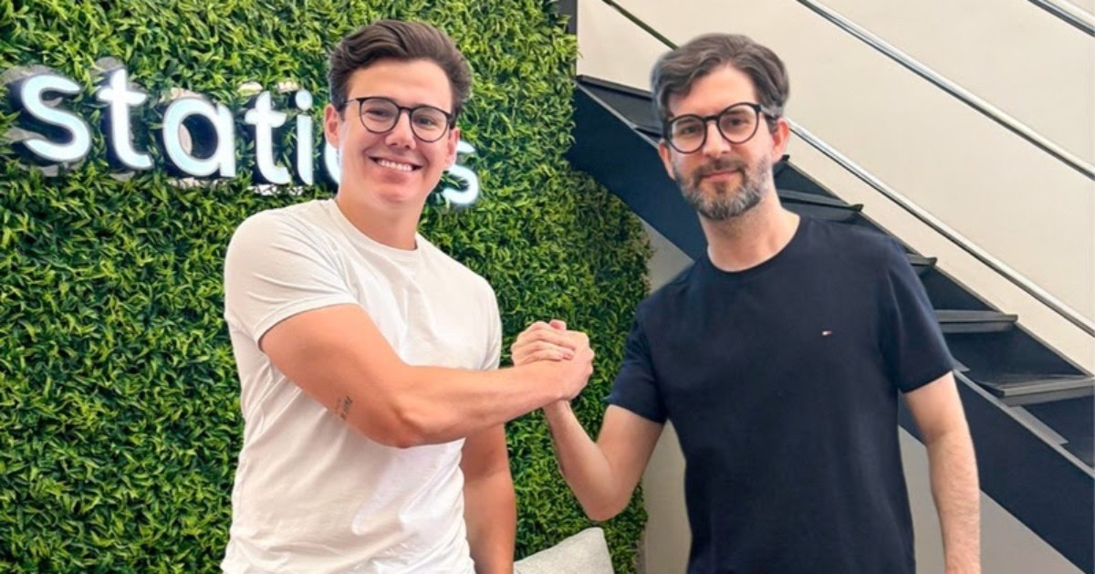 Dois homens, um de camisa branca e outro de camisa preta, ambos de óculos, posando para uma foto