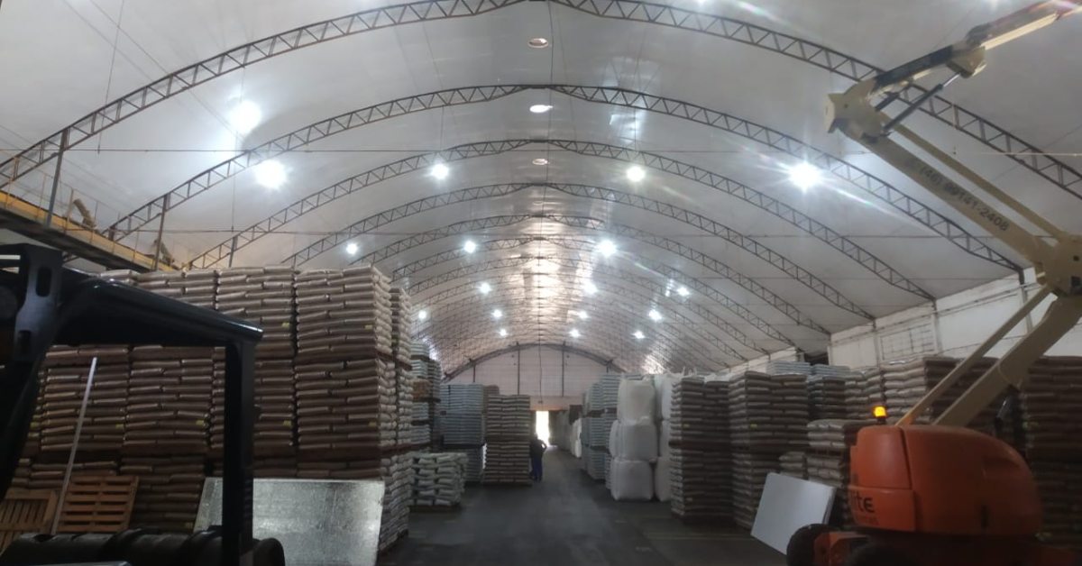 A imagem mostra o interior de um grande galpão industrial, com teto alto e arqueado, sustentado por estruturas metálicas. O espaço é bem iluminado por várias lâmpadas instaladas ao longo do teto.