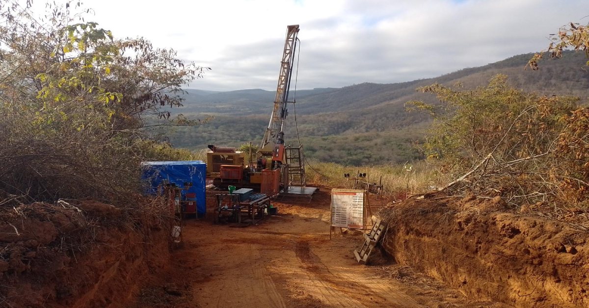 Um local de perfuração ao ar livre com uma plataforma de perfuração amarela alta cercada por arbustos e colinas arborizadas sob um céu nublado. Na frente, há uma caixa azul, equipamentos e placas em uma estrada de terra.