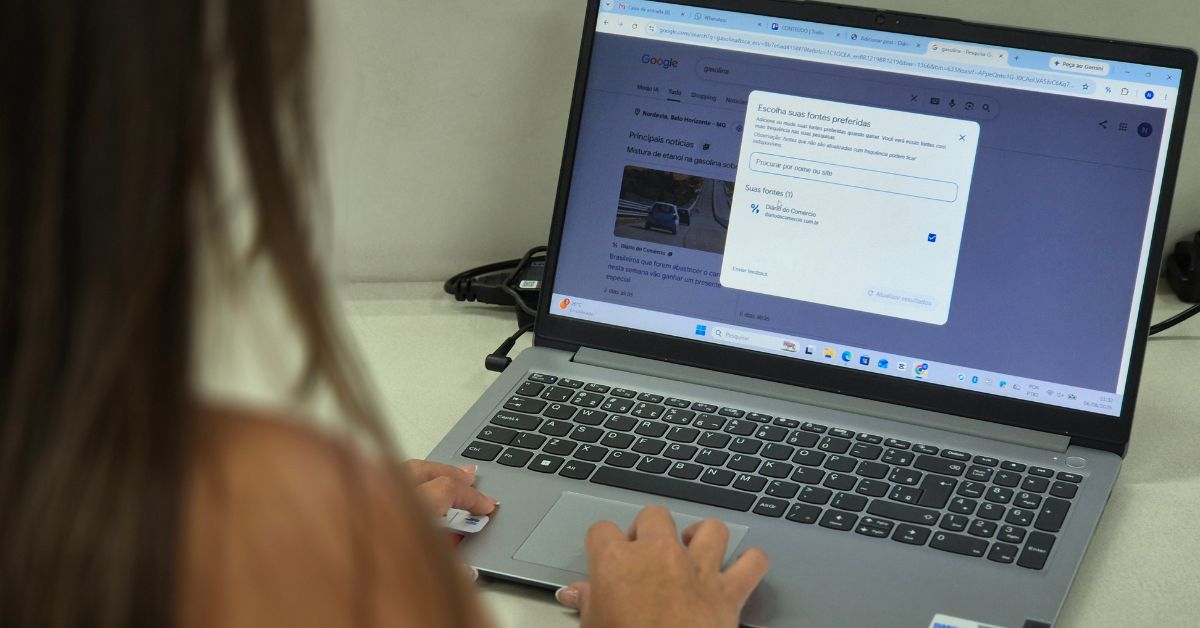 Mulher utiliza um notebook para acessar o Google Notícias enquanto a janela "Escolha suas fontes preferidas" está aberta, com o Diário do Comércio selecionado como uma das fontes favoritas. A imagem ilustra o processo de personalização das preferências de notícias na plataforma.