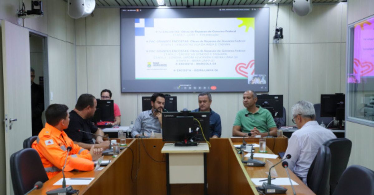 Reunião em sala da Câmara Municipal de Belo Horizonte, com participantes sentados ao redor de mesas e uma apresentação projetada em uma tela ao fundo.