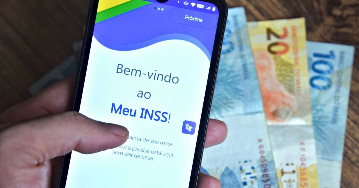 Mão segurando um celular com o aplicativo Meu INSS aberto e notas de dinheiro ao fundo.
