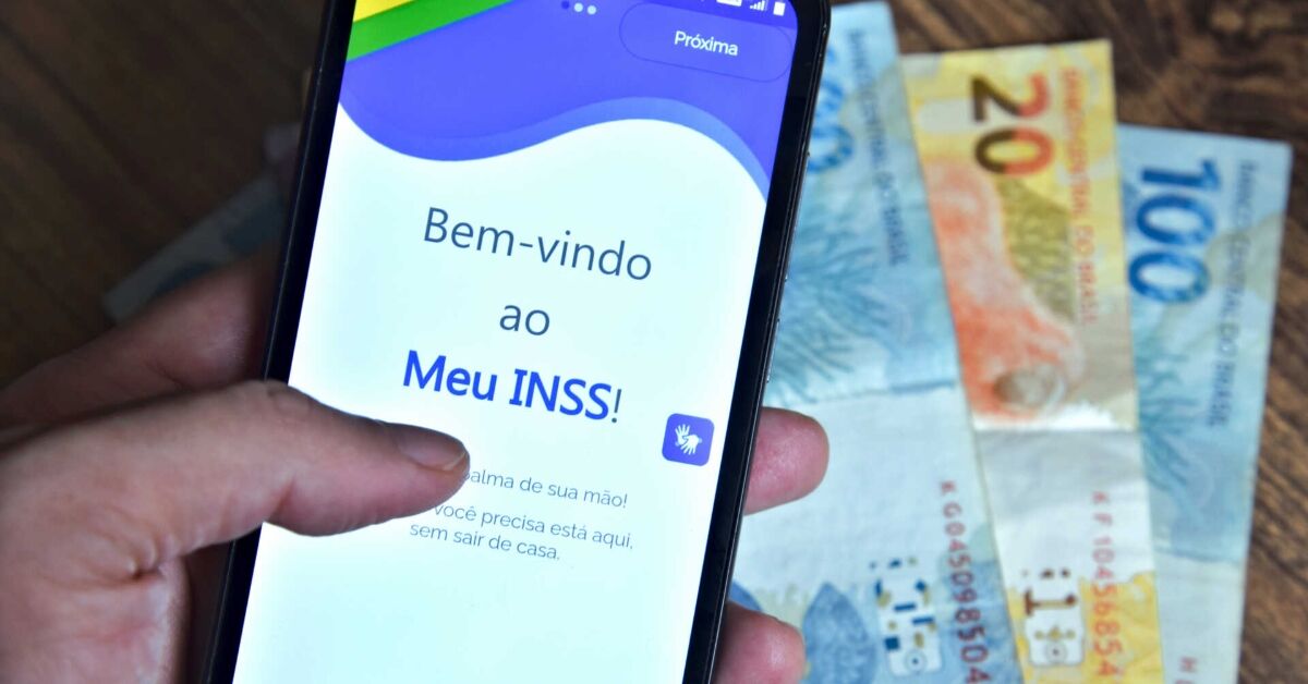 Mão segurando um celular com o aplicativo Meu INSS aberto e notas de dinheiro ao fundo.