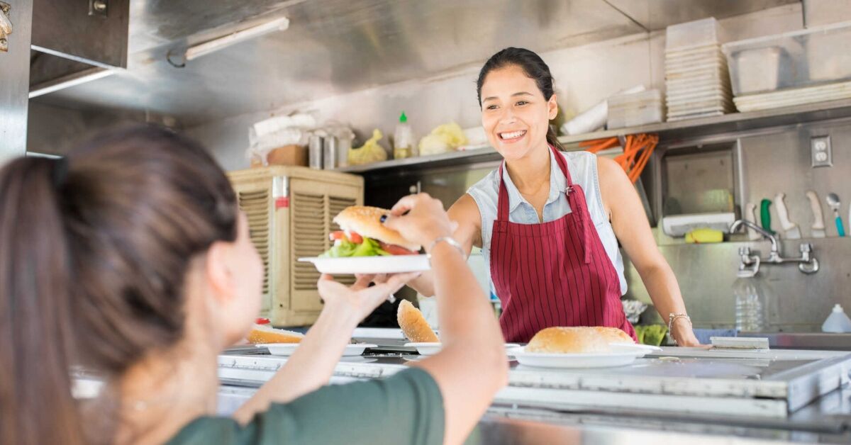 Funcionária sorridente de food truck entregando um sanduíche para uma cliente.
