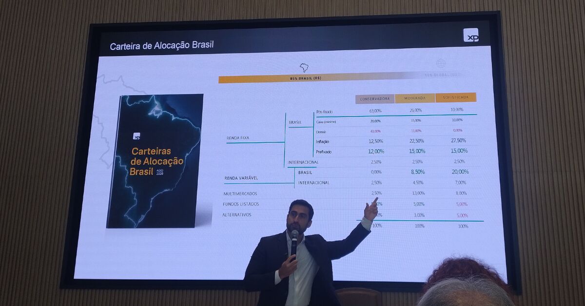 Homem de terno apresentando em um evento corporativo com o logotipo da XP ao fundo.