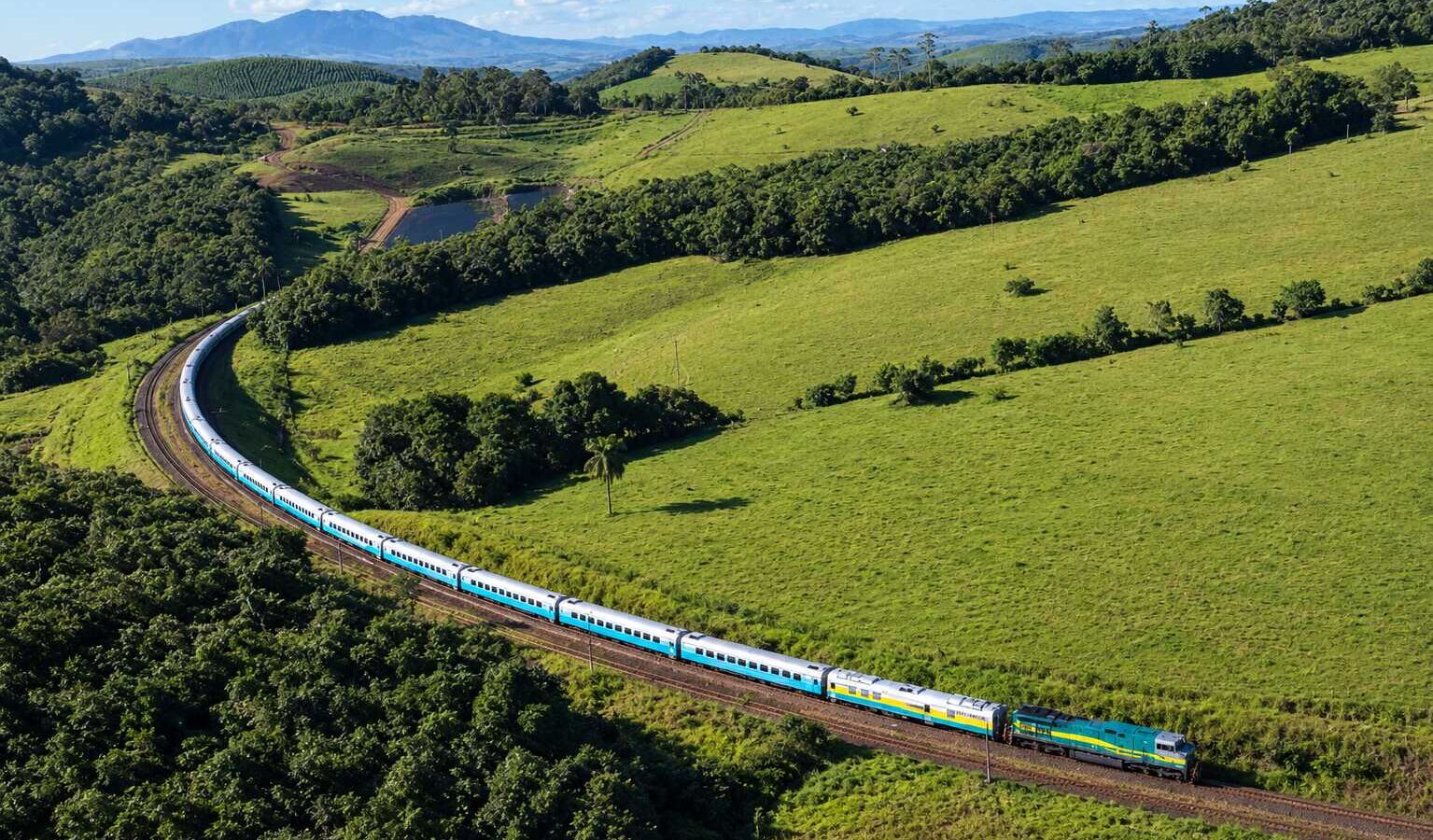 Trem de passageiros atravessando uma paisagem rural com campos verdes e florestas.