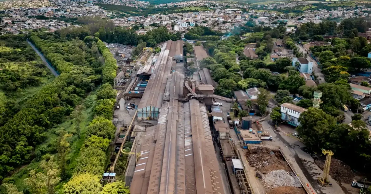 Fotografia aérea de um vasto complexo industrial com longos galpões metálicos e pátios de materiais, ladeado por uma densa vegetação e com uma cidade ao fundo sob um céu nublado.