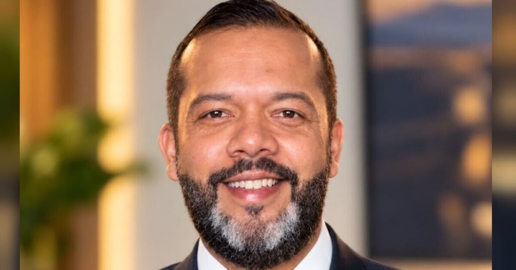 Homem sorrindo, de barba grisalha e cabelo curto, vestindo terno azul-marinho, camisa branca e gravata bordô, em ambiente interno com iluminação suave.